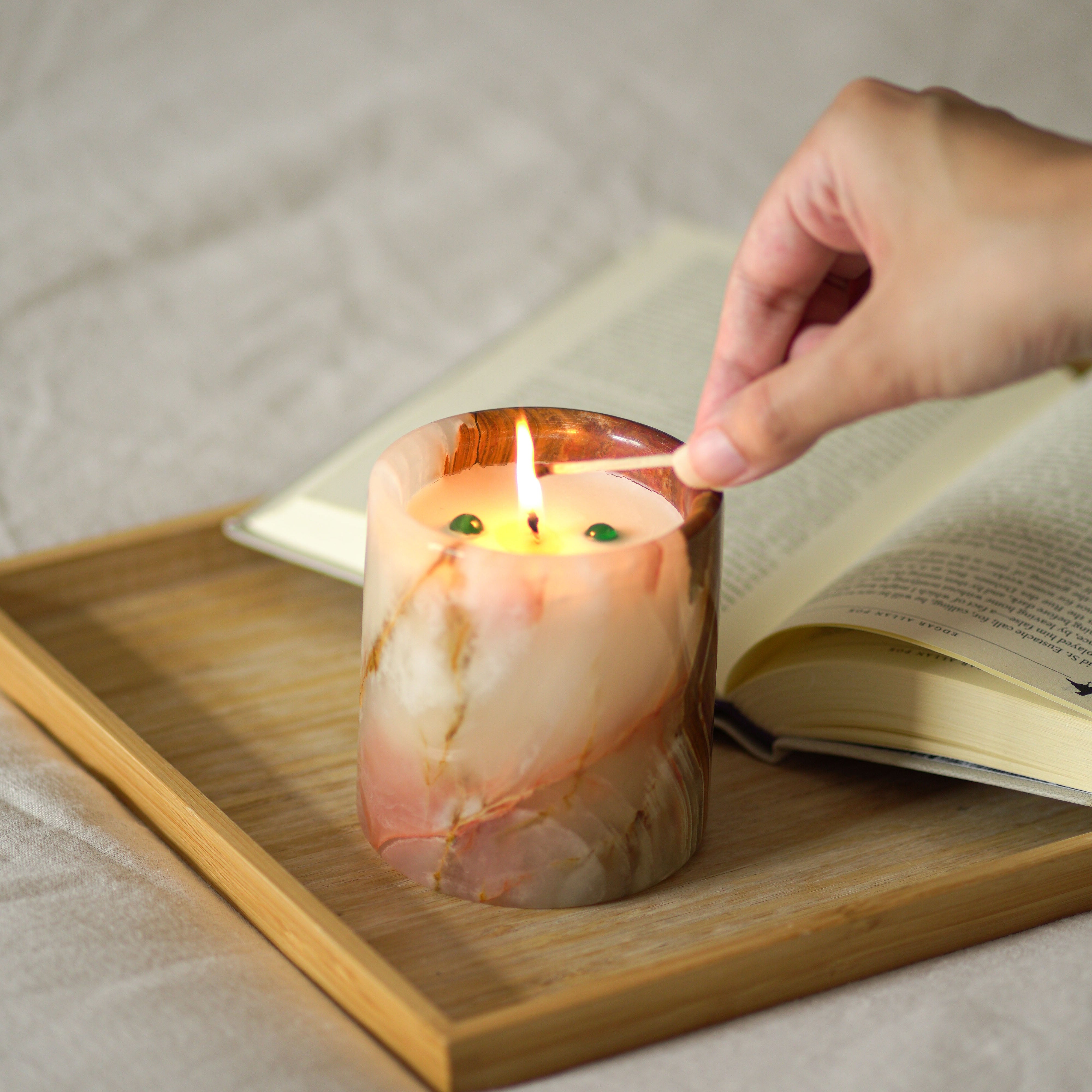 Hand lighting a candle in a marbled container on a wooden tray with a book underneath.